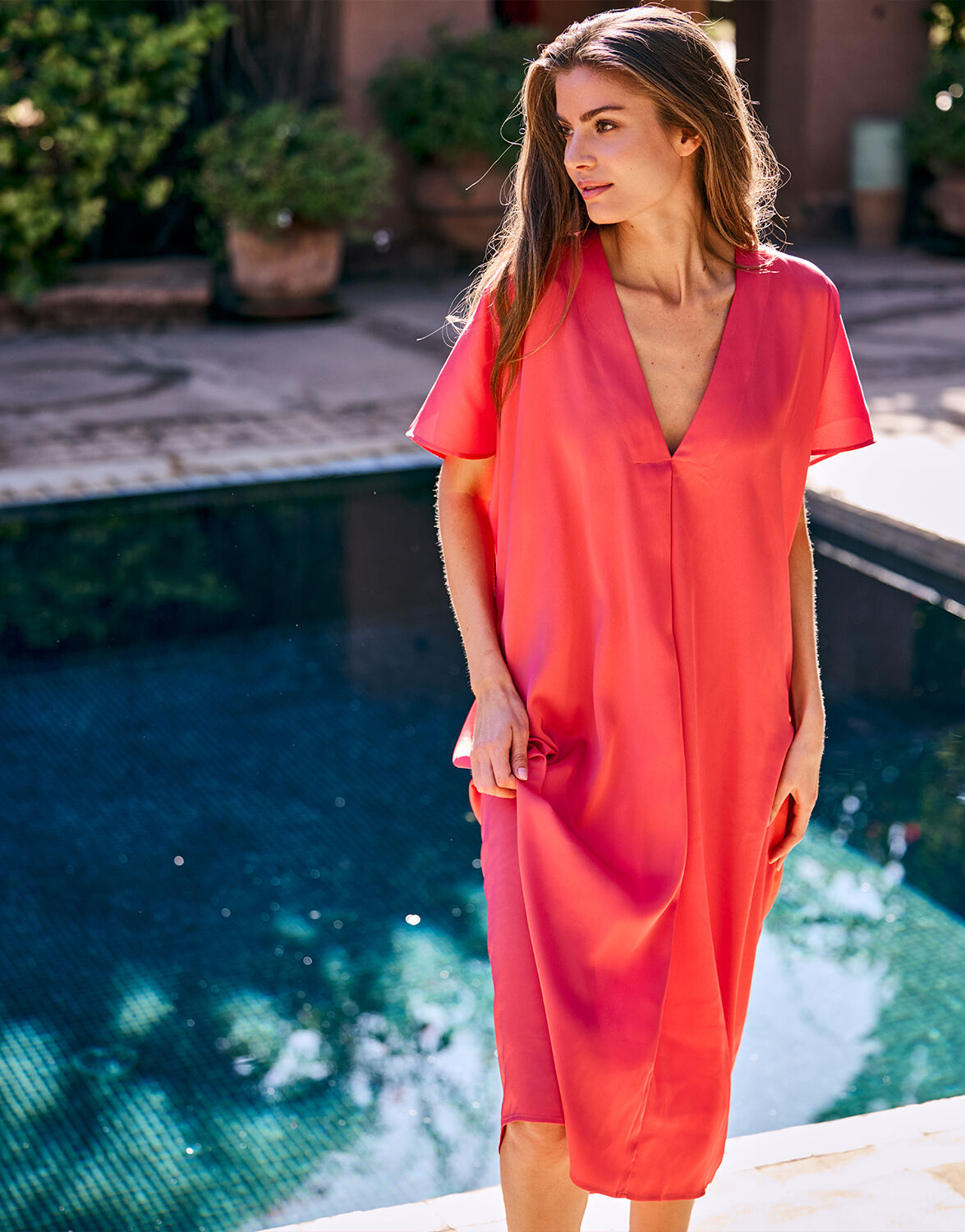 Model standing in the Ilaria Coral dress in satin with a V-neck and kaftan shape.