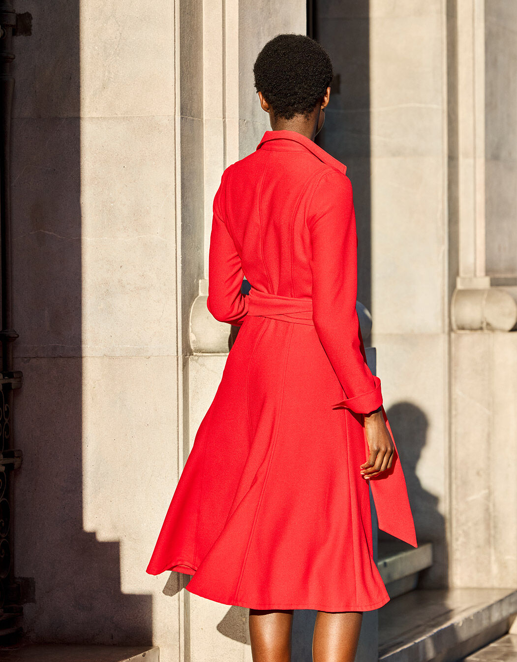 Red wool coat worn by a person in front of a marble facade in sunlight, elegant and stylish design of the fashionable overcoat.