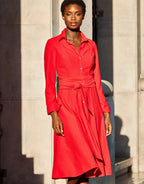 Red dress with button detail and long sleeves in sunlight against a classic stone wall, elegant and timeless design.