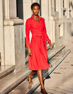 Woman in a stylish red dress with a belt walks past a classic stone building in a sunny urban setting.