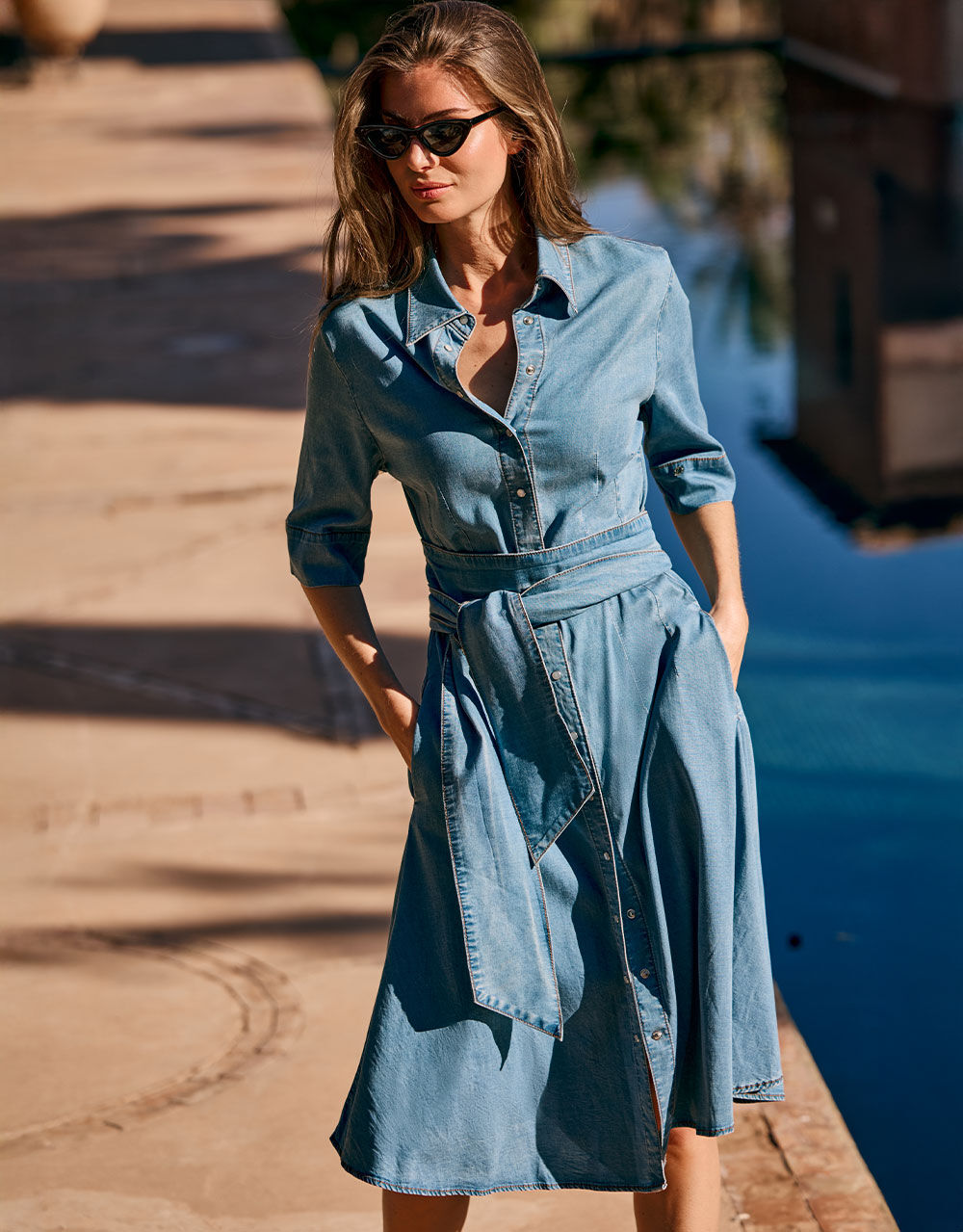 Long blue denim dress with button closure and tie belt, worn by a woman with sunglasses and medium-length hair.
