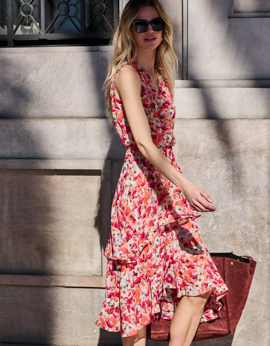 Woman in an elegant pink floral dress on a sunny day, with long wavy hair and black sunglasses.