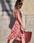 Woman in colorful floral dress with ruffles walks into a wall outside, wearing stylish sunglasses and carrying a brown bag.