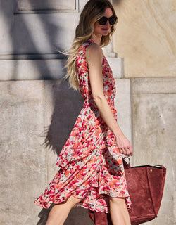 Woman in colorful floral dress with ruffles walks into a wall outside, wearing stylish sunglasses and carrying a brown bag.