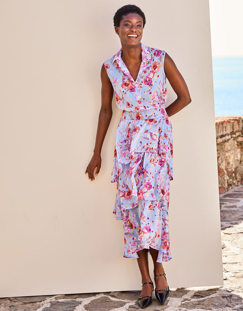 Frau in einem bunten Blumenkleid mit plissierten Lagen und Knöpfen, stehend vor einer beigefarbenen Wand an einem sonnigen Tag.