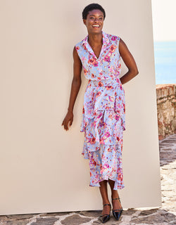 Woman in a colorful, floral dress with pleated layers and buttons, standing against a beige wall on a sunny day.