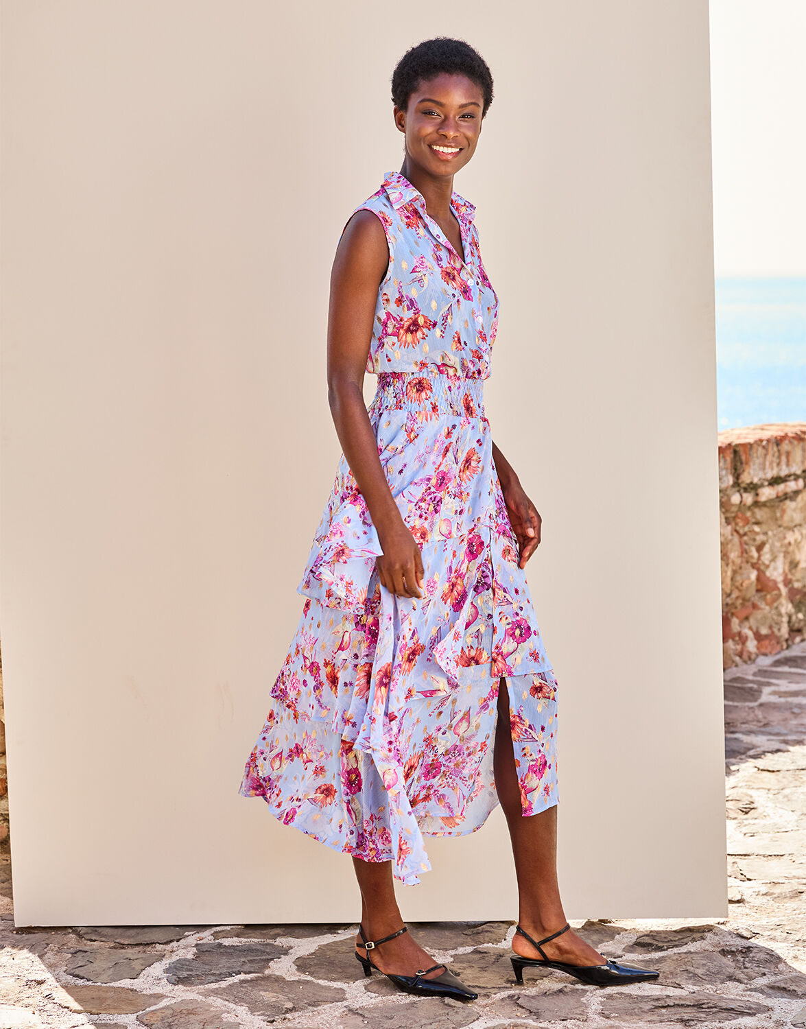 Woman in a summery sleeveless floral dress with ruffles, standing on a sunny day with a smile on her face.