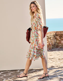 Woman in an elegant, floral summer dress with ruffles, walking outside with a brown shoulder bag and sandals.