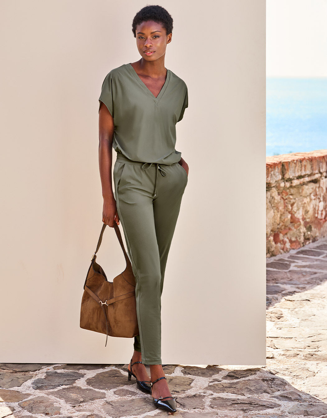 Model standing in the Valencia Olive trousers in jersey lycra with a suede bag.
