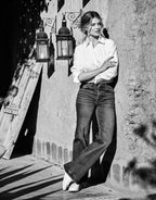 Model wears white blouse and jeans, leaning against a rough stone wall with decorative lanterns.