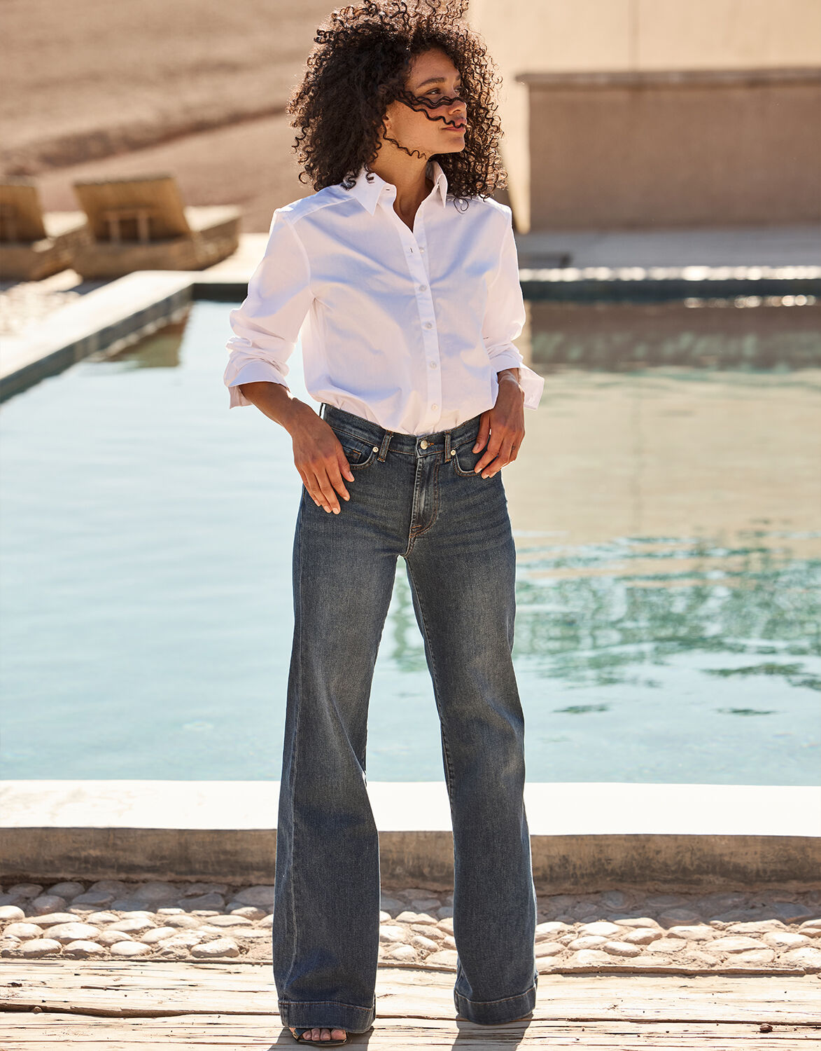 White long-sleeved blouse and straight, dark blue jeans, worn by a model in front of a modern swimming pool.