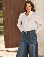 Woman in a white blouse and dark blue jeans in a sunny outdoor setting with a wooden background.