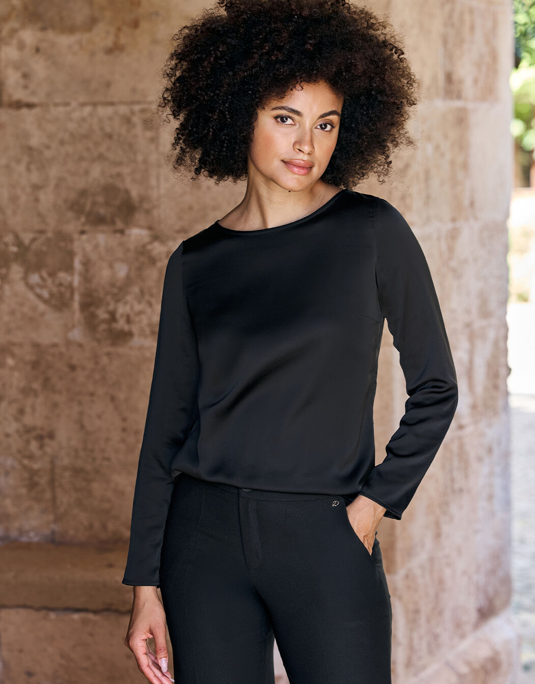 Model standing in the Dorian Black top in satin with a boat neck and long sleeves.