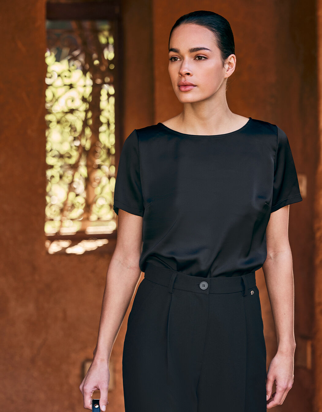 Model standing in the Monica Black top in satin with a round neck and short sleeves.