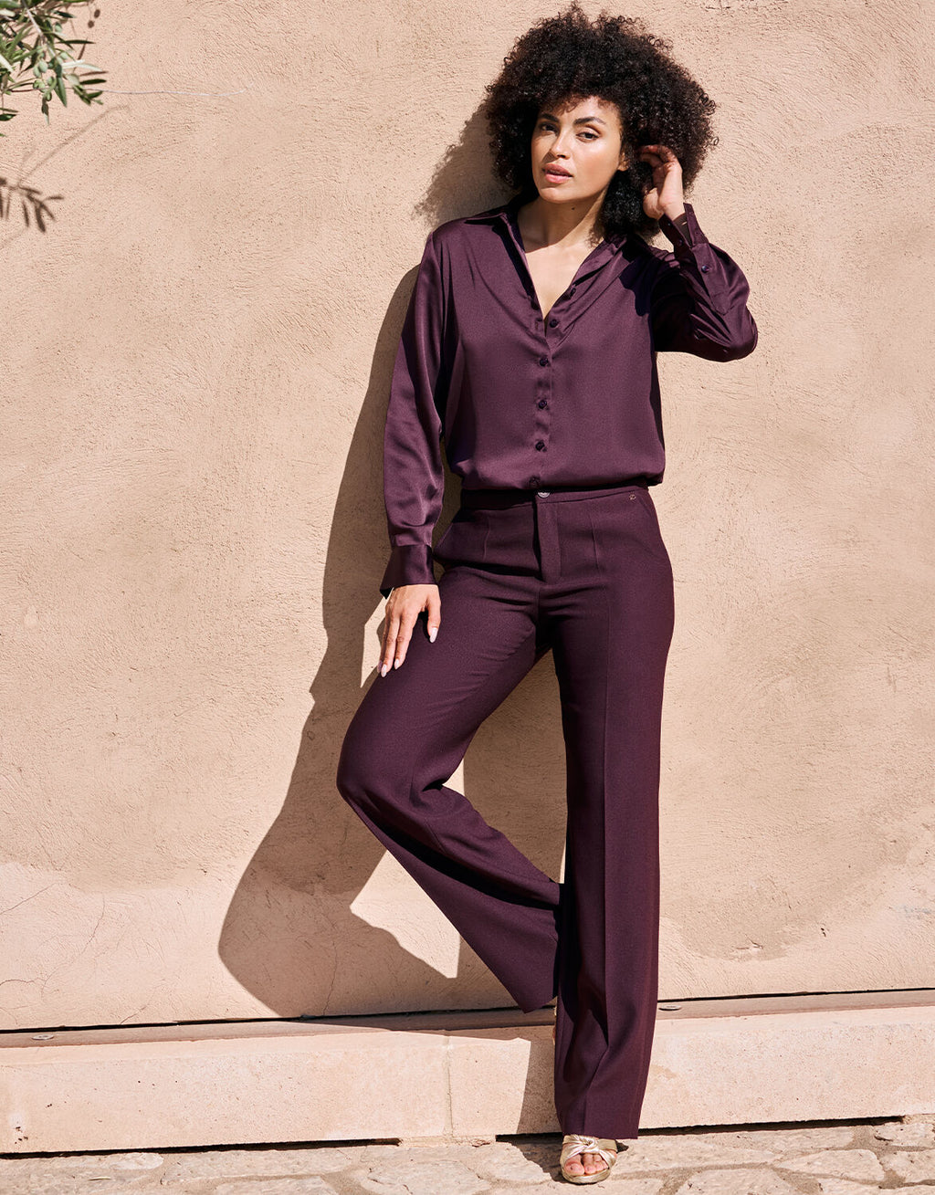 Elegant woman poses in a dark purple silk blouse and trousers against a beige plastered wall in the sun.