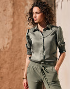 Green satin blouse with long sleeves and smocked details, worn by a woman outdoors, against a brown-beige wall.