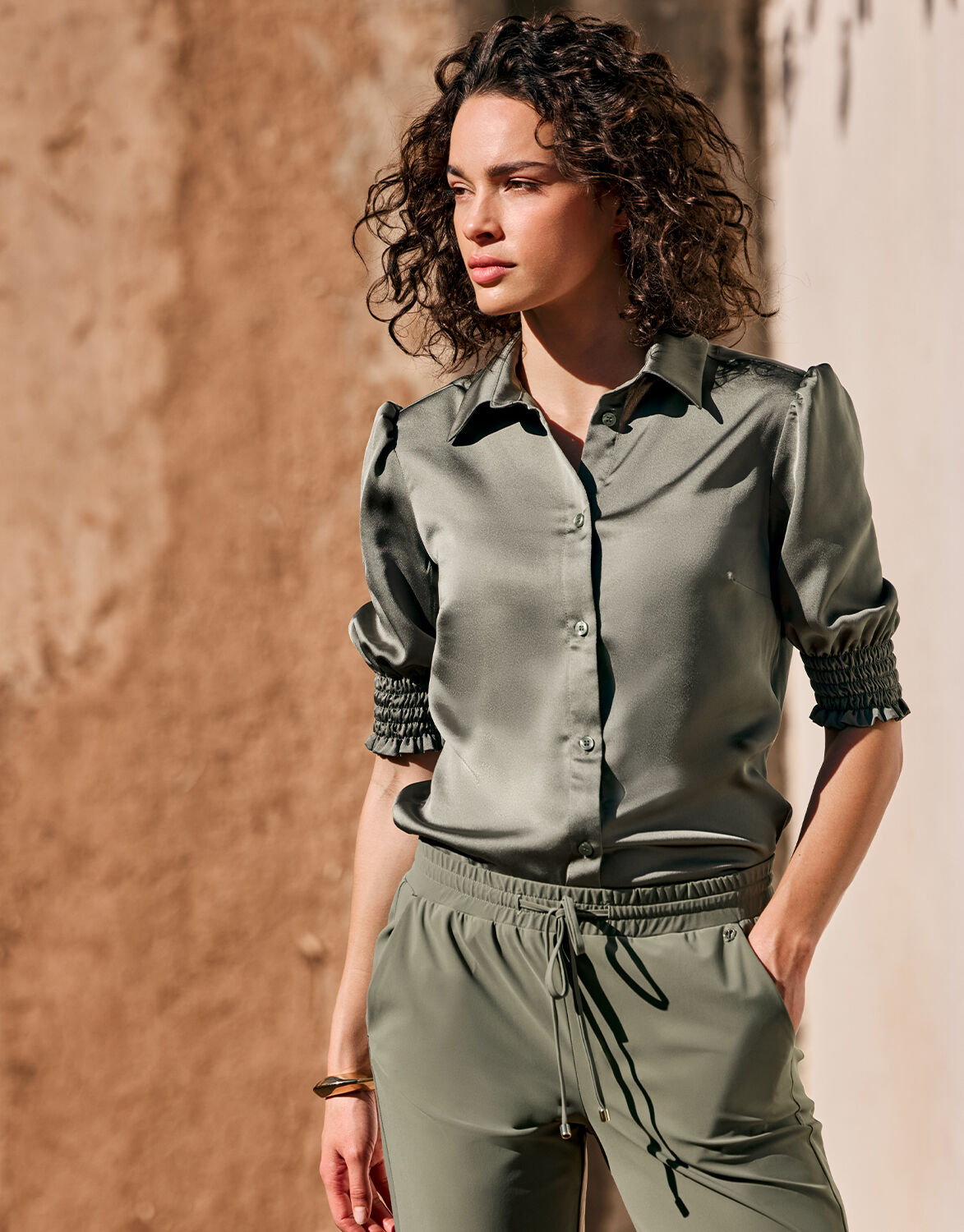 Green satin blouse with long sleeves and smocked details, worn by a woman outdoors, against a brown-beige wall.