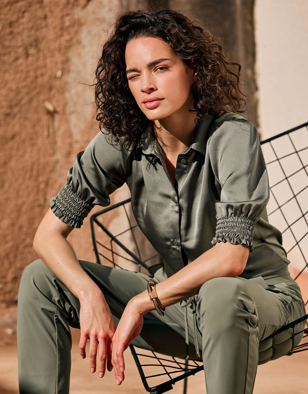 Woman in a green satin blouse with pleated sleeves, sitting on a metal wire chair in a sunny setting.