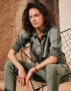 Woman in a green satin blouse with pleated sleeves, sitting on a metal wire chair in a sunny setting.