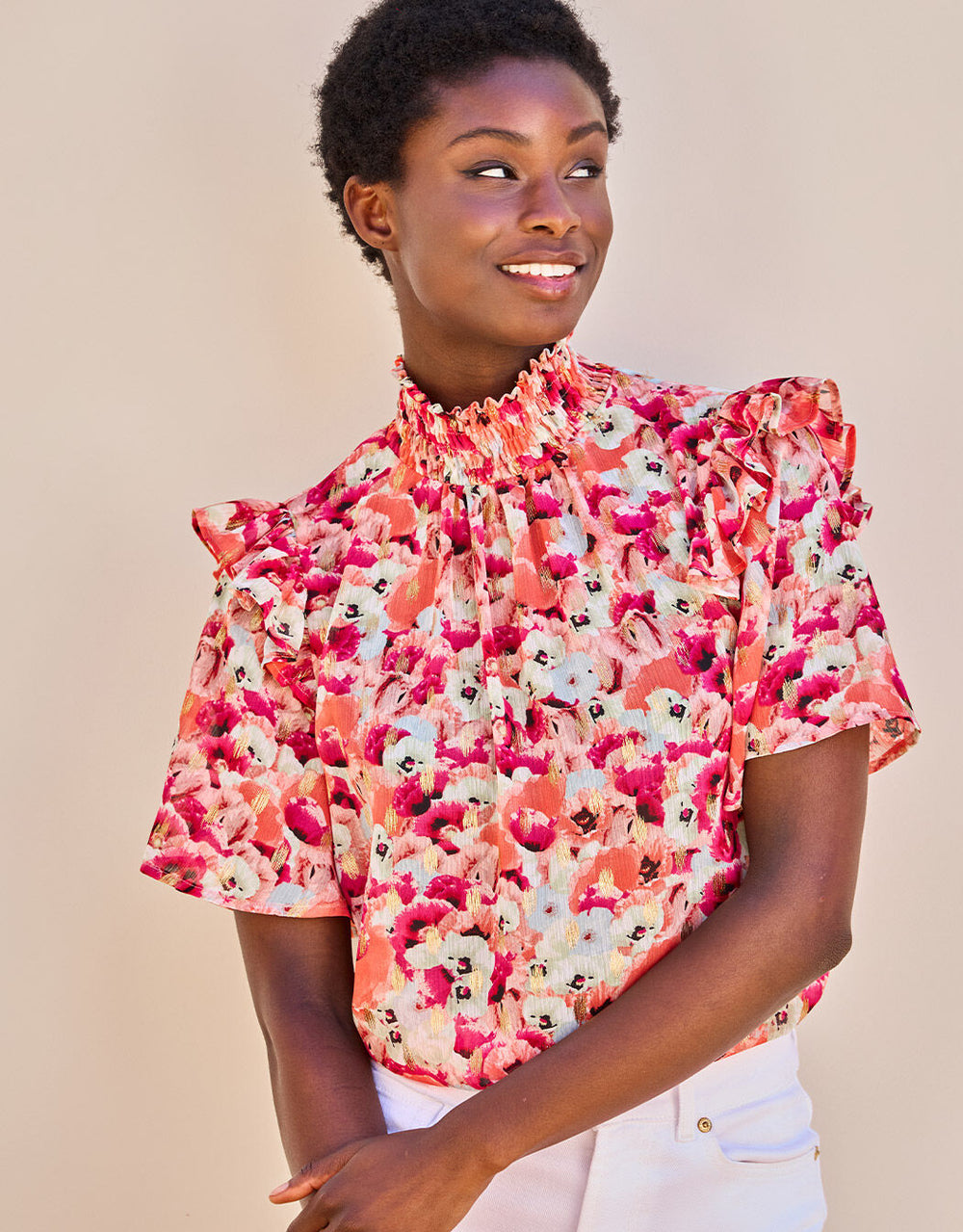 Frau in einer bunten Bluse mit Blumenprint, Rüschen und hohem Kragen, stehend vor neutralem Hintergrund.