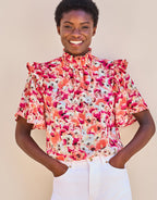 Felix blouse with pink floral print and ruffled collar, worn by a person with hands in white trouser pockets.