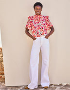 Woman in a colorful blouse with a floral print and white wide-leg pants, leaning against a neutral wall.