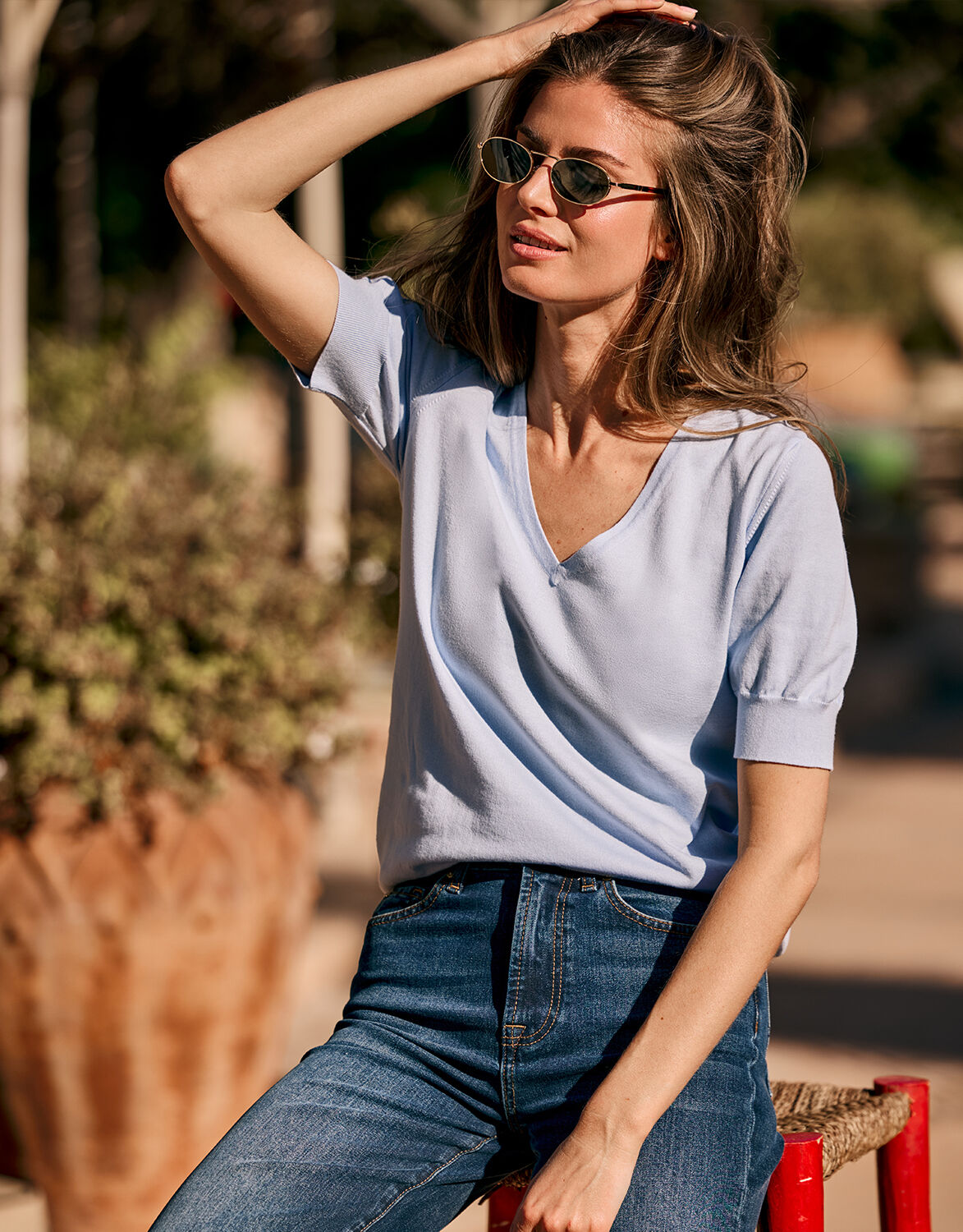 Vrouw in casual zomeroutfit met lichtblauwe V-hals T-shirt en donkerblauwe jeans poserende in de zon.