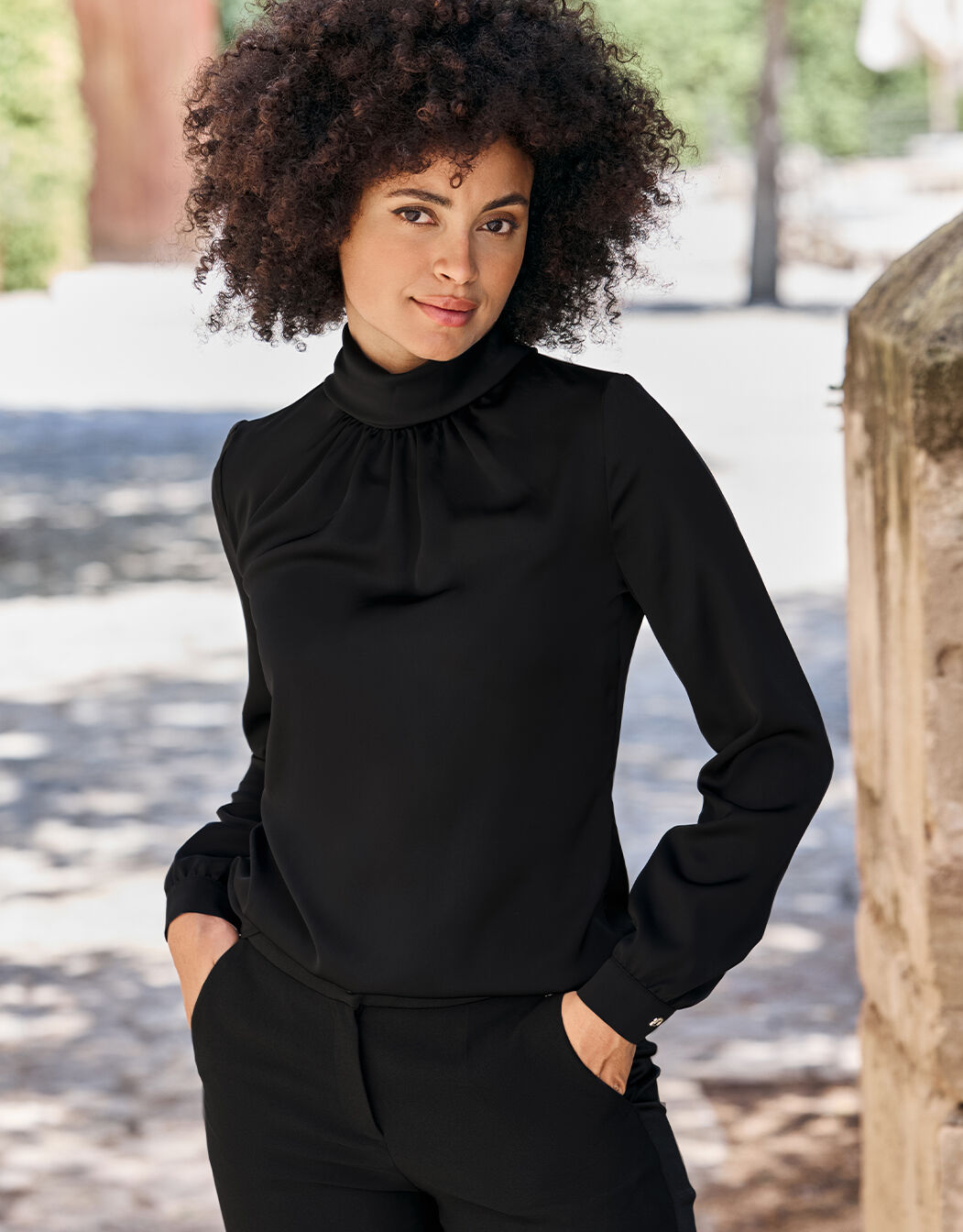 Model standing in the Vittoria Black blouse in satin with a gathered high neck.