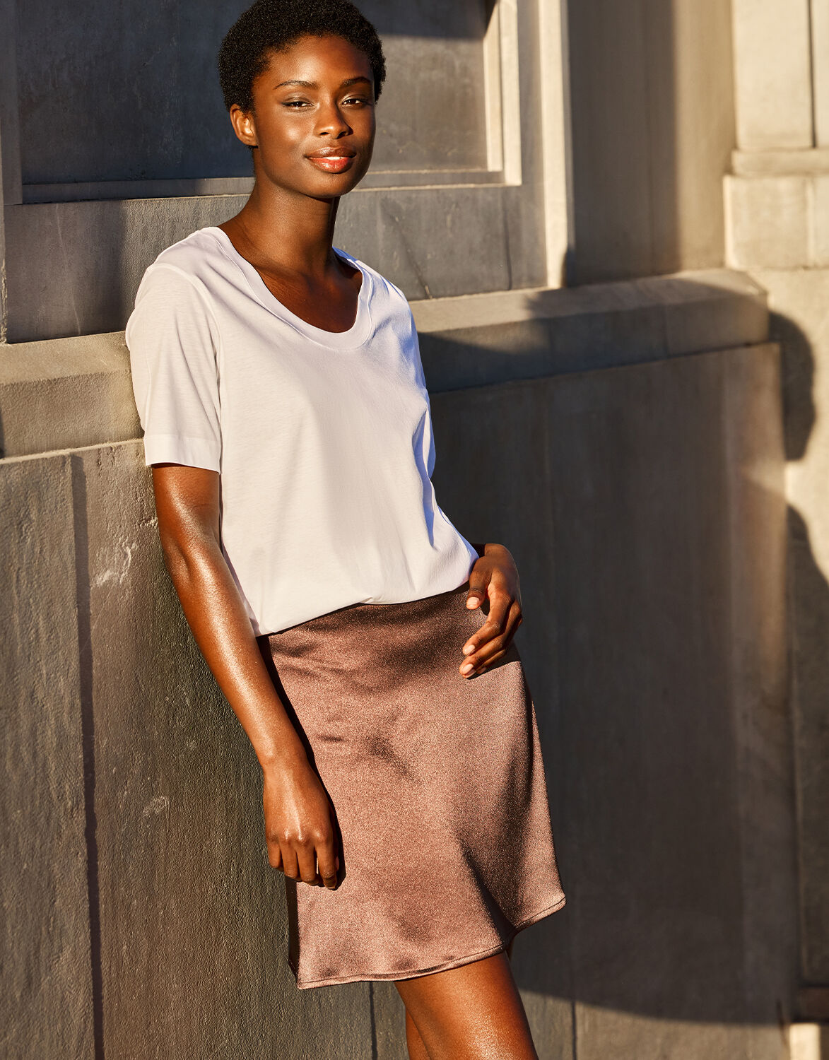 Vrouw draagt een witte, korte mouw t-shirt en een glanzende, bruine rok, leunend tegen een betonnen muur in zonlicht.