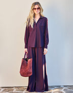 Woman in a stylish purple blouse and wide-legged trousers, paired with a burgundy suede bag and sunglasses.