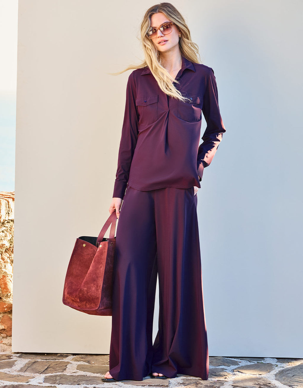 Female model in an elegant, burgundy silk blouse and trousers, wears dark sunglasses and carries a red suede bag.