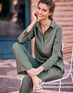 Woman in an olive green pantsuit with rolled-up sleeves, sitting on a white metal chair, smiles at the camera.
