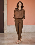 Brown silk blouse and earth-toned trousers for an elegant style, worn by a woman on a marble floor with a peach-colored wall.