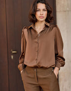 Woman in a silky brown blouse with long sleeves and an elegant collar, accentuated by a wooden door in the background.