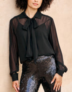 Black transparent blouse with bow detail, worn with shimmering black trousers, against a beige background.