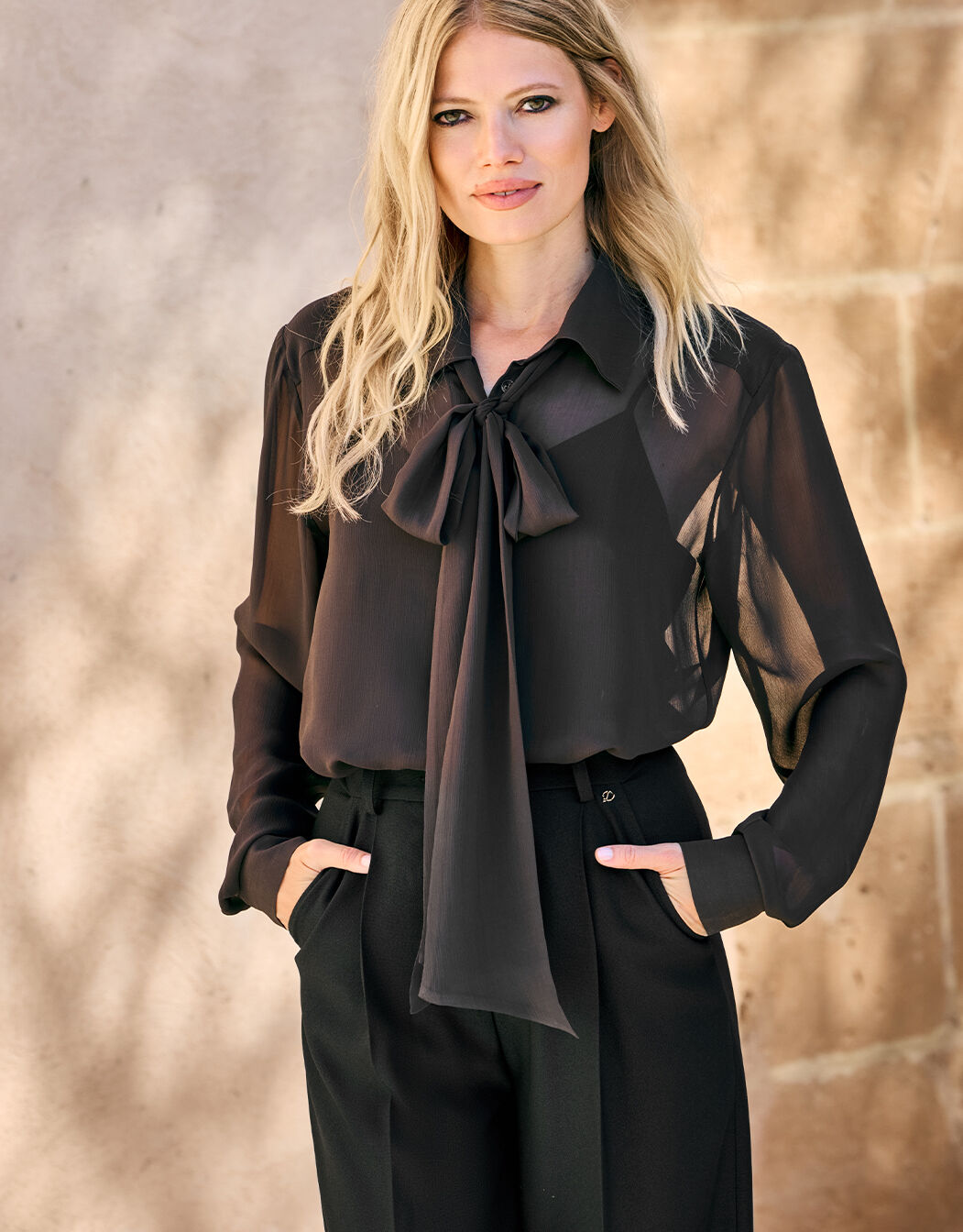Model standing in the Nola Black blouse in chiffon with a pussy bow and black trousers.