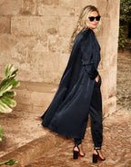 Woman in a stylish dark blue satin trench coat with sunglasses, standing by a stone wall in an outdoor setting.