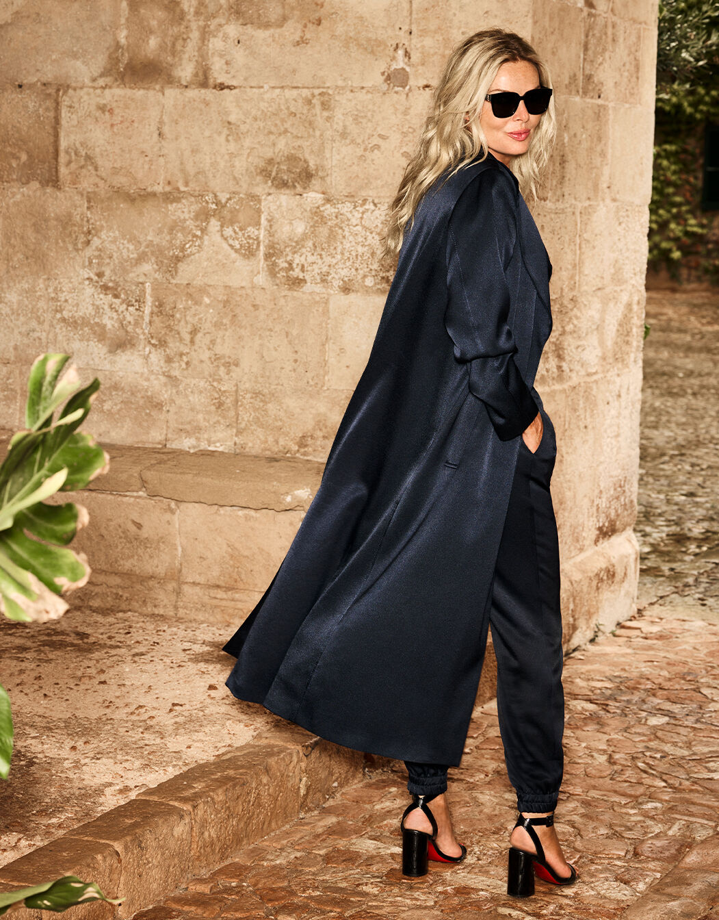 Vrouw in stijlvolle donkerblauwe satijnen trenchcoat met zonnebril, staand bij een stenen muur in een buitenomgeving.