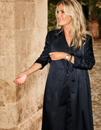 Woman in an elegant long black coat against a rustic stone wall, with a stylish wristwatch.