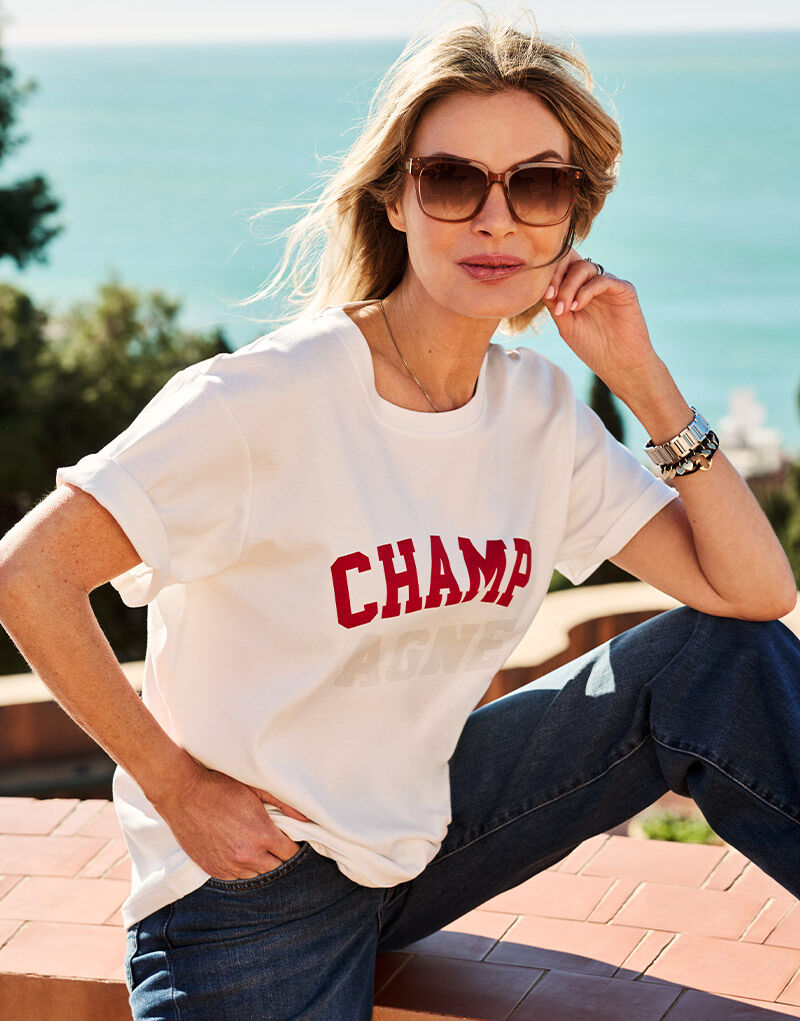 Model seated in the Story Tee Off White Champ in stretch cotton with a CHAMPAGNE print.