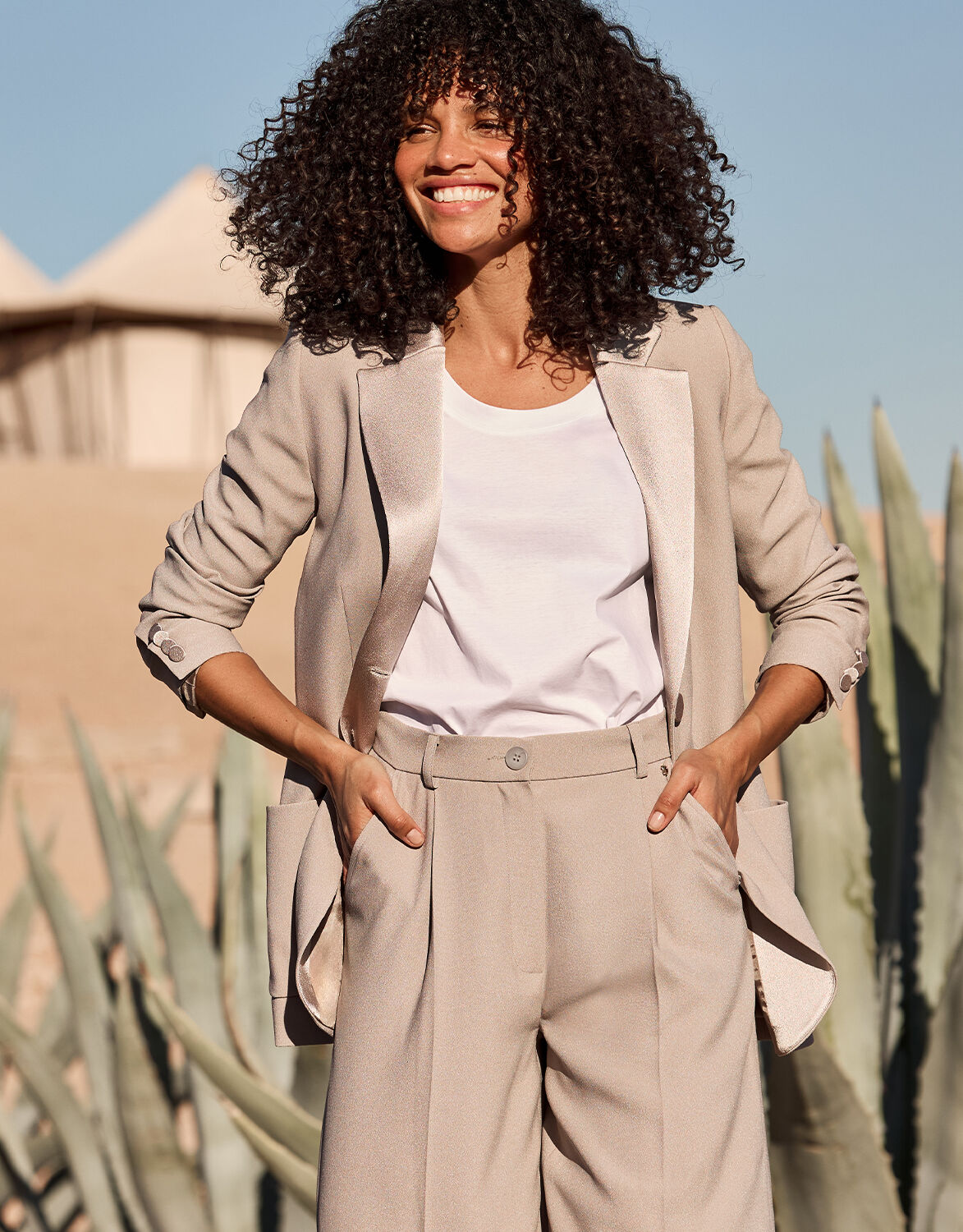 Model standing in the Bianca Pebble blazer in crepe satin, worn open over a white top, hands in trouser pockets.