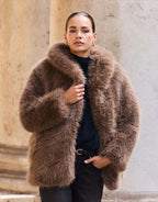 Model standing in the Isolde Dark Taupe coat in faux fur, holding the collar.