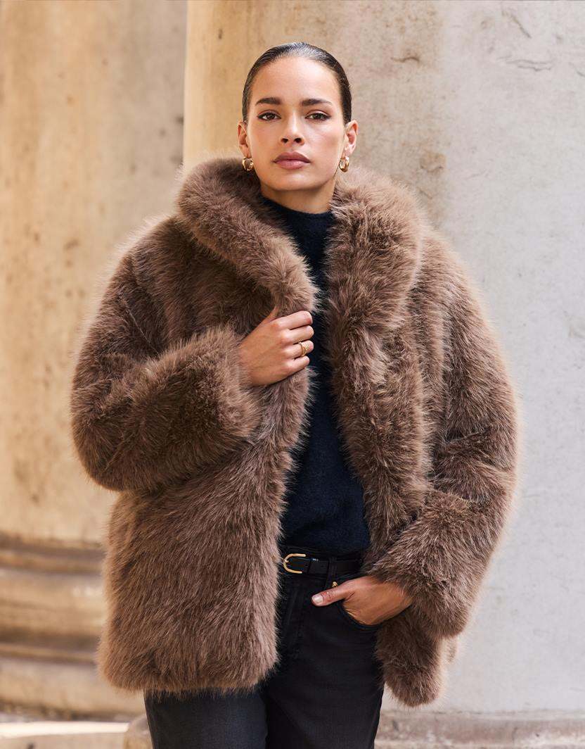 Model standing in the Isolde Dark Taupe coat in faux fur, holding the collar.