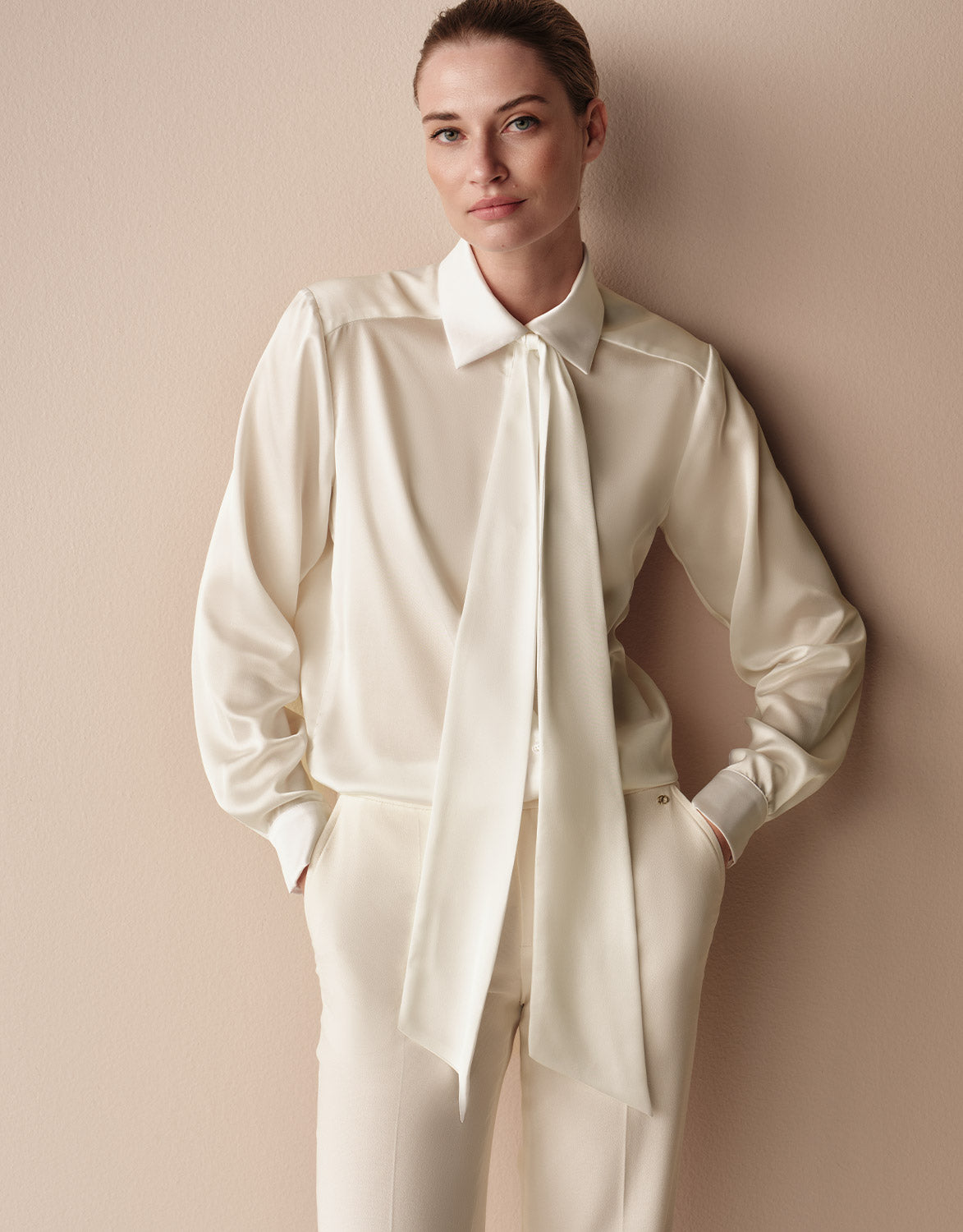 Model wearing the Addy Ivory satin blouse with a tie neck and matching ivory trousers.