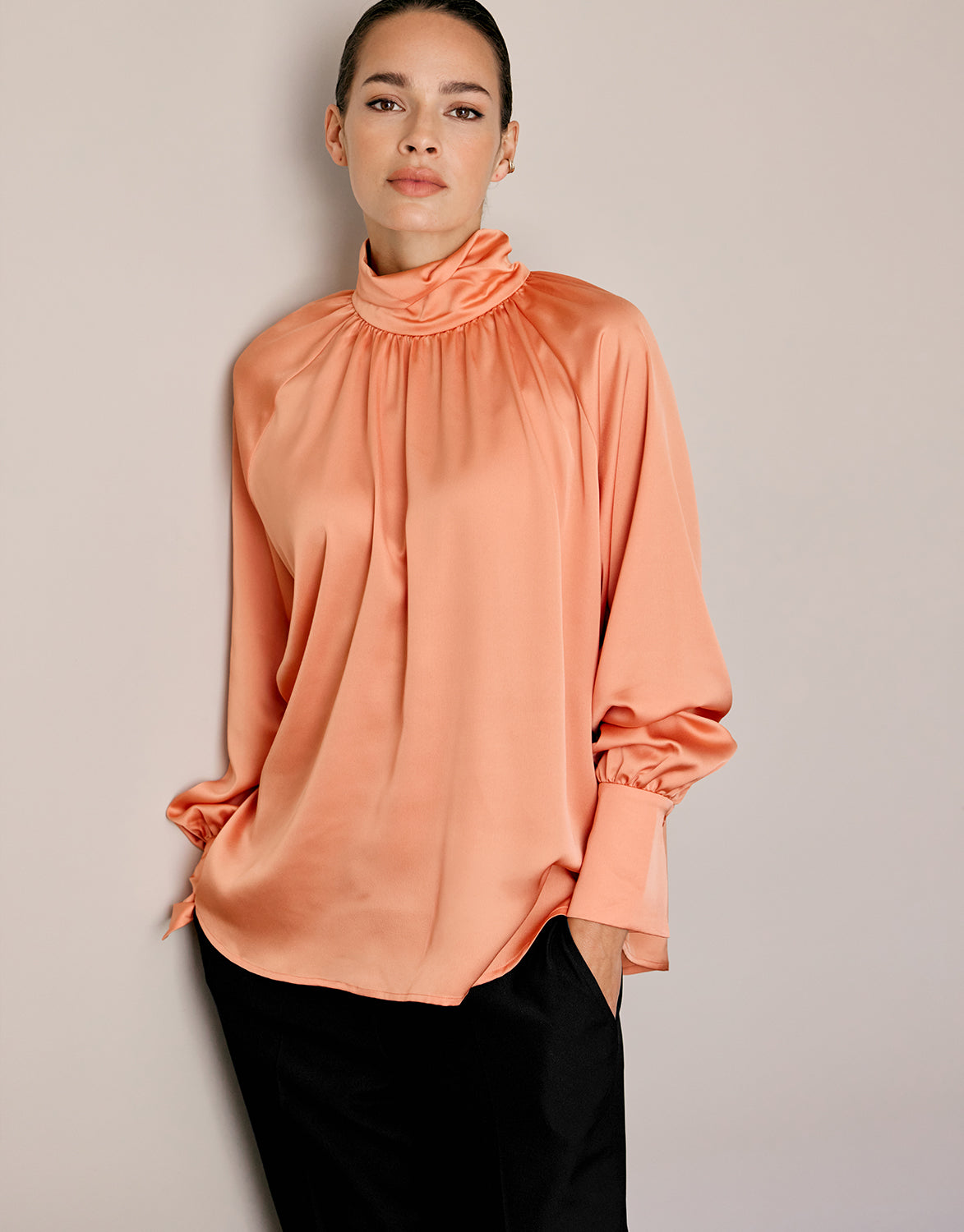 Model wearing the Alix Soft Apricot satin blouse with a high neck and black trousers.