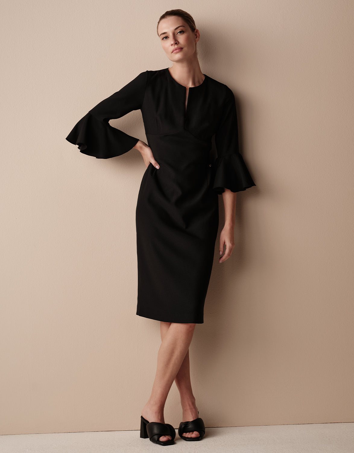 Model standing in the Daphne Black dress in crepe satin with flared flounce sleeves.