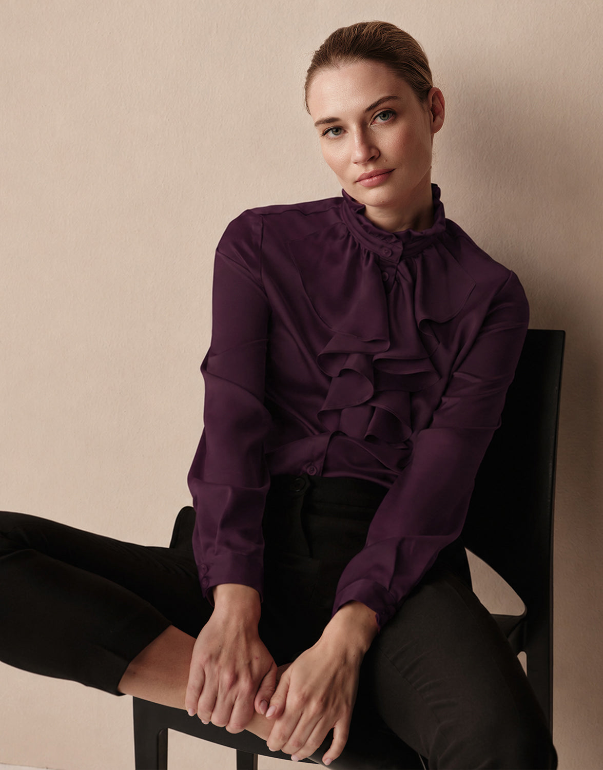 Model seated in the Eloise Deep Aubergine blouse in satin with a ruffled jabot front.