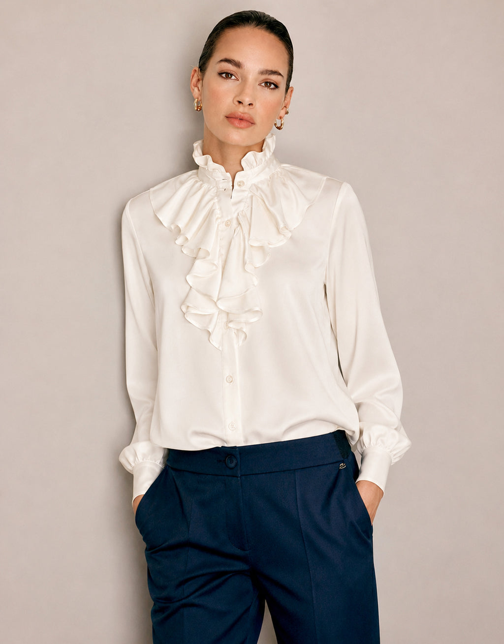 Model steht in der Eloise Ivory Bluse aus Satin mit Volantvorderseite und marineblauer Hose.