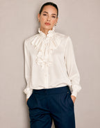 Model steht in der Eloise Ivory Bluse aus Satin mit Volantvorderseite und marineblauer Hose.