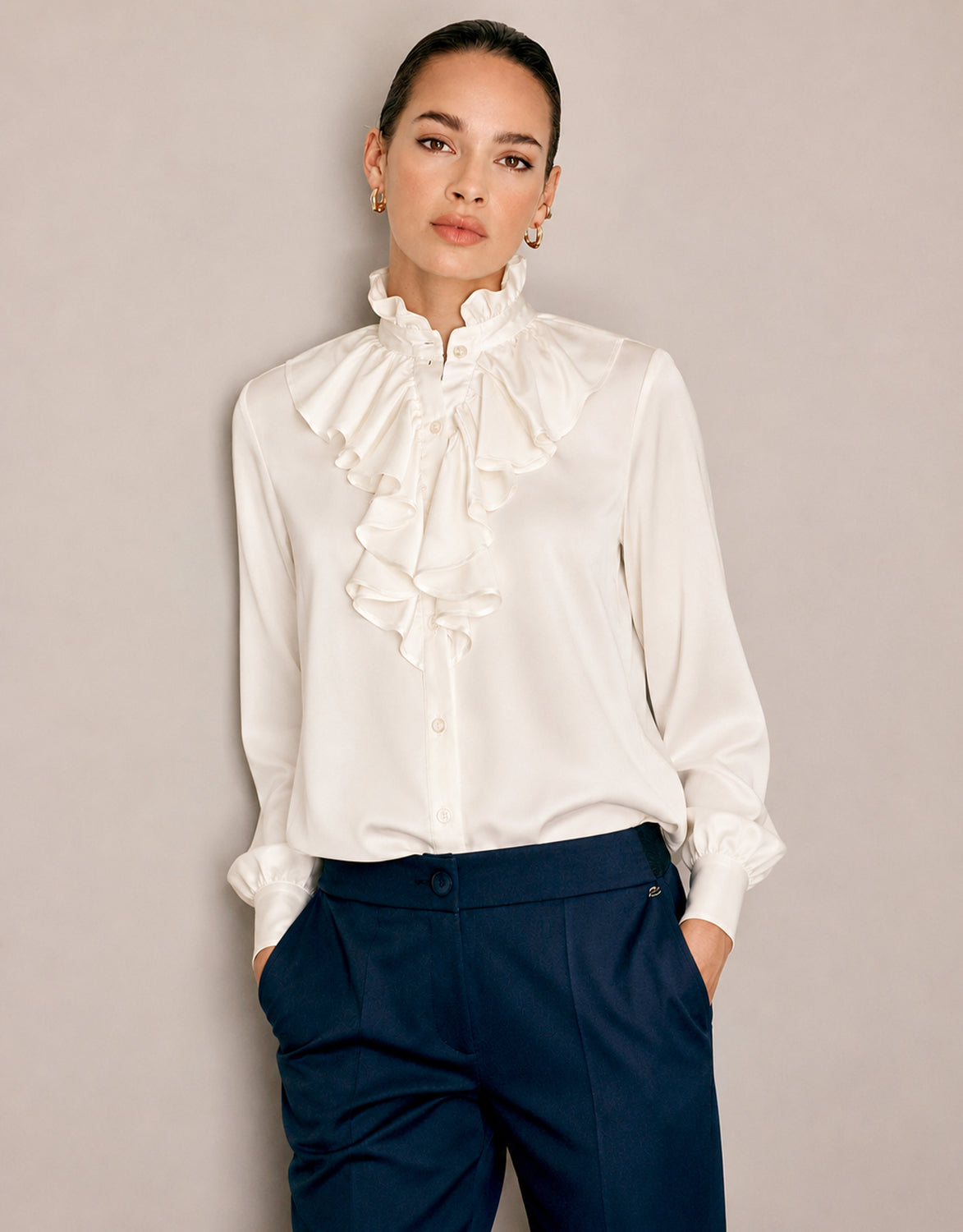 Model steht in der Eloise Ivory Bluse aus Satin mit Volantvorderseite und marineblauer Hose.